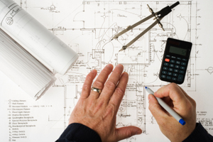 Man updating blueprints and performing project estimates using a calculator and geometric compass.